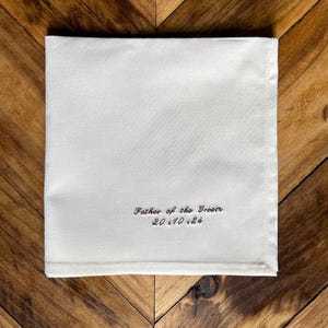 May include: A cream-coloured square handkerchief with the embroidered text "Father of the Groom" and the date "20.10.24" in brown thread. The handkerchief is placed on a wooden surface.