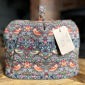 Puede incluir: Un cubreteiera acolchado con un colorido estampado floral y de pájaros en rojo, naranja, verde y azul. El cubreteiera tiene un asa de lazo y una etiqueta con el texto "Handmade with Love" y un logotipo.