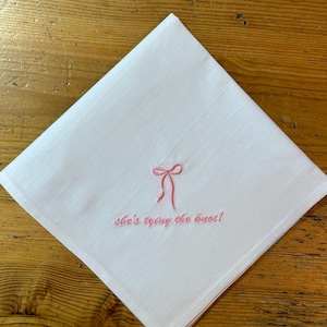Könnte beinhalten: Weiße Leinenserviette mit rosa Stickerei eines Bogens und dem Text "She's tying the knot!"
