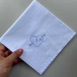 以下が含まれることがあります： 中央に青い刺繍が施された正方形の白いハンカチ。デザインは矢が貫通したハートの形で、ハートの内側にはLとAの文字が縫い込まれています。生地は滑らかな質感で、縁は丁寧に仕上げられています。