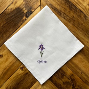 May include: White handkerchief featuring an embroidered purple iris flower and the name "Sylvia". The fabric is folded and set on a wooden surface with a chevron pattern.