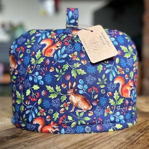 Puede incluir: Un calentador de té azul marino con un estampado de bosque con ardillas, conejos, bayas y follaje. El calentador tiene un asa de bucle y una etiqueta adjunta. El diseño de la tela incluye elementos rojos, verdes y azules.