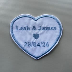 Può includere: Una toppa ricamata a forma di cuore bianco con bordo blu. La toppa presenta i nomi "Leah & James", un piccolo cuore e la data "28/04/26" ricamati con filo blu. Ideale per un matrimonio o un anniversario.