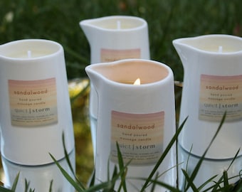 May include: Four white ceramic massage candles with a pitcher design, labeled "sandalwood quiet | storm." One candle is lit, showing a warm glow. The candles are set on a reflective surface with green grass in the background.