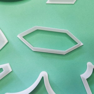 May include: A set of white plastic cookie cutters in various shapes, including a hexagon, a crescent moon, and a rectangle with rounded corners. The cutters are arranged on a light green surface.