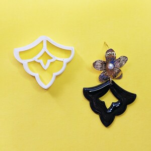 Könnte beinhalten: Ein weißer Ausstecher in dekorativer Form liegt neben einem goldfarbenen und schwarzen Ohrring mit Blumenmuster. Der Ohrring hat eine Perle in der Mitte der Blume. Der Hintergrund ist gelb.