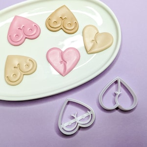 Puede incluir: Galletas en forma de corazón en tonos rosa y beige, algunas con caras sonrientes y otras con un diseño dividido, dispuestas sobre un plato ovalado blanco. También se ven dos cortadores de galletas en forma de corazón, lo que sugiere un tema de repostería.