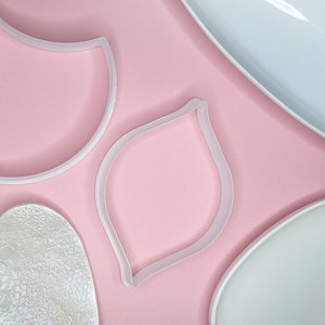 Puede incluir: Tres cortadores de galletas de plástico blanco sobre un fondo rosa. Los cortadores tienen forma de corazón, hoja y media luna.