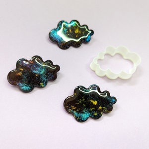 Puede incluir: Cuatro objetos con forma de nube sobre una superficie de color lila claro. Tres son brillantes, con una base oscura y motas coloridas de azul, dorado y verde. También hay un cortador de galletas blanco con forma de nube.