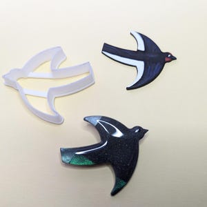 May include: Three cookie cutters in the shape of a bird. One cutter is white plastic, and the other two are painted black and green.