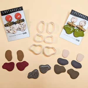 May include: A set of six clay earrings in various colors and patterns, including green, brown, red, and white. The earrings are displayed on a light beige surface with two cards that say "Lostangela Handmade".