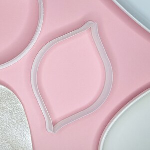 May include: Four white plastic cookie cutters on a pink background. The cutters are shaped like a circle, a leaf, and two different sized ovals.