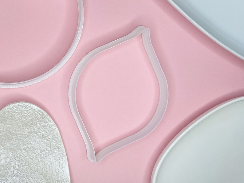 May include: Four white plastic cookie cutters on a pink background. The cutters are shaped like a circle, a leaf, and two different sized ovals.