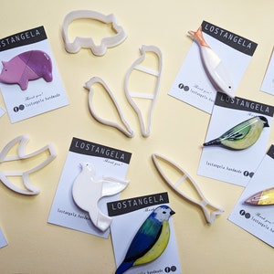May include: A collection of handmade ceramic animal brooches, including a pig, a bird, a fish, and a bear. The brooches are displayed on white cards with the brand name "LOSTANGELA" and the text "Thank you! lostangela handmade".