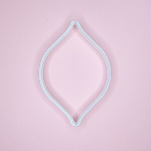 May include: White plastic cookie cutter shaped like a teardrop or a leaf. The cutter is on a pink background.