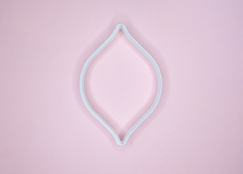 May include: White plastic cookie cutter shaped like a teardrop or a leaf. The cutter is on a pink background.
