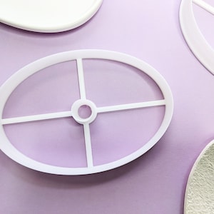 Può includere: Tagliapasta ovale in plastica bianca con un design interno a forma di croce. Il tagliapasta è su una superficie lilla chiaro, con altri oggetti bianchi sullo sfondo. Per la cottura e l'artigianato.