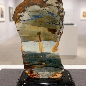 May include: A polished, vertically oriented slab of multicoloured jasper stone featuring horizontal bands of tan, sage green, deep blue, and rust orange, mounted securely on a glossy black rectangular display base.