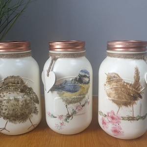 May include: Three white painted mason jars with copper lids and twine accents. Each jar features a different bird illustration: a robin, a blue tit, and a wren. Each jar has a small white heart attached to the twine.