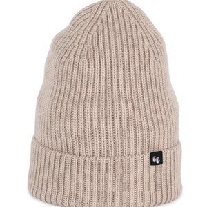 A dark sand-beige beanie with chunky, fisherman-style ribbed knit. It has a black fabric label with a white logo stitched to the cuff