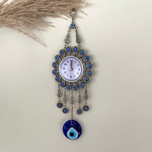 May include: A silver clock with a white face and blue evil eye charms hanging from it. The clock is surrounded by a circle of blue evil eyes. The clock is hanging from a chain with a large blue evil eye charm at the bottom.