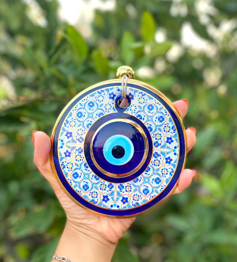May include: A round, blue and white ceramic evil eye wall hanging with a gold trim. The evil eye is surrounded by a white background with blue and gold floral patterns.