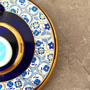 May include: A blue and white ceramic lid with a gold rim and a blue evil eye design. The lid is decorated with a white and gold floral pattern.