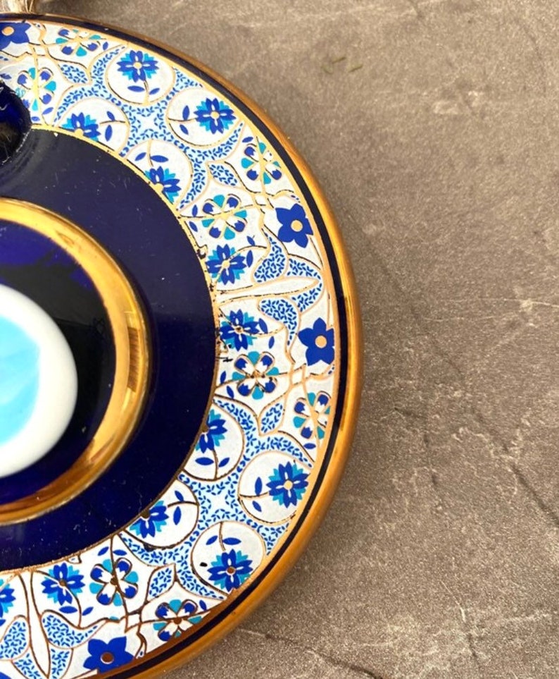May include: A blue and white ceramic lid with a gold rim and a blue evil eye design. The lid is decorated with a white and gold floral pattern.