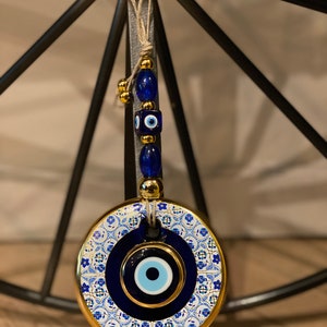 May include: A round, blue and gold evil eye charm with a white and blue patterned design. The charm is hanging from a string of blue and gold beads.
