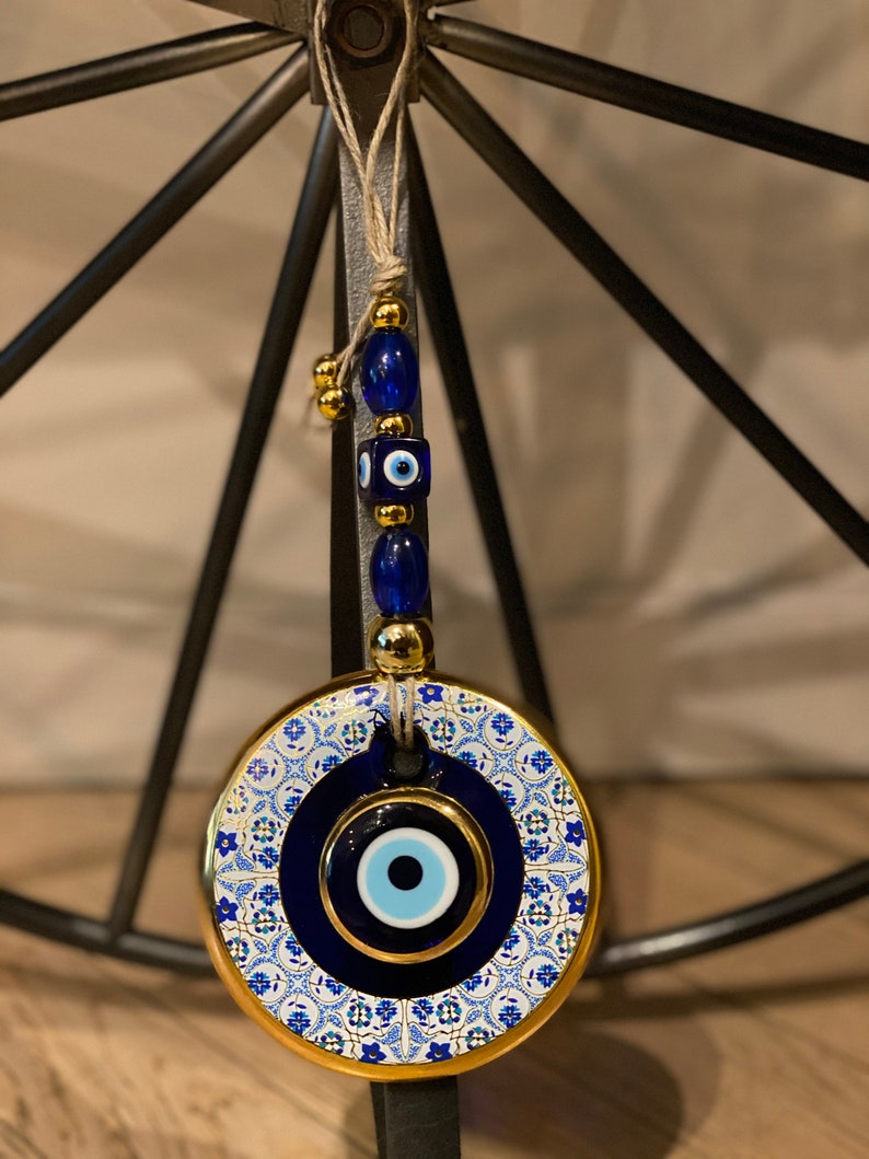 May include: A round, blue and gold evil eye charm with a white and blue patterned design. The charm is hanging from a string of blue and gold beads.
