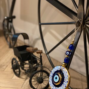 May include: A blue and white ceramic evil eye charm with gold accents hanging from a black metal wheel. The charm is round and has a blue eye in the center. The wheel is decorated with a white and blue pattern.