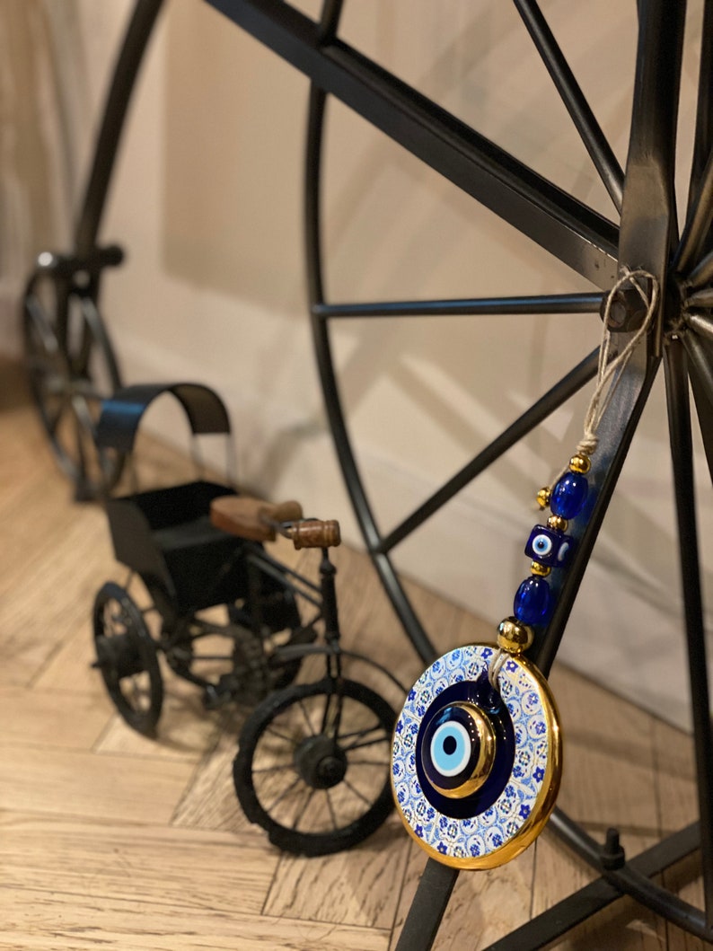 May include: A blue and white ceramic evil eye charm with gold accents hanging from a black metal wheel. The charm is round and has a blue eye in the center. The wheel is decorated with a white and blue pattern.