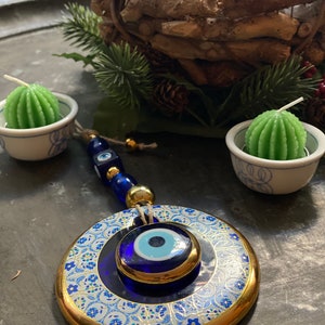 May include: A blue and gold evil eye charm with a white background and intricate floral patterns. The charm is hanging from a string of blue and gold beads.