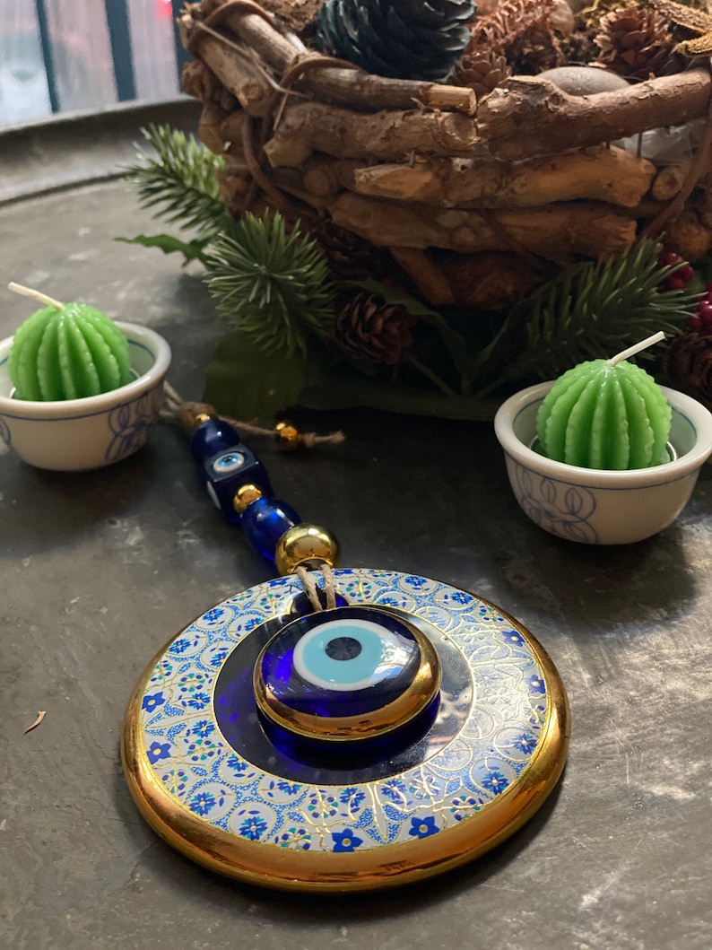 May include: A blue and gold evil eye charm with a white background and intricate floral patterns. The charm is hanging from a string of blue and gold beads.