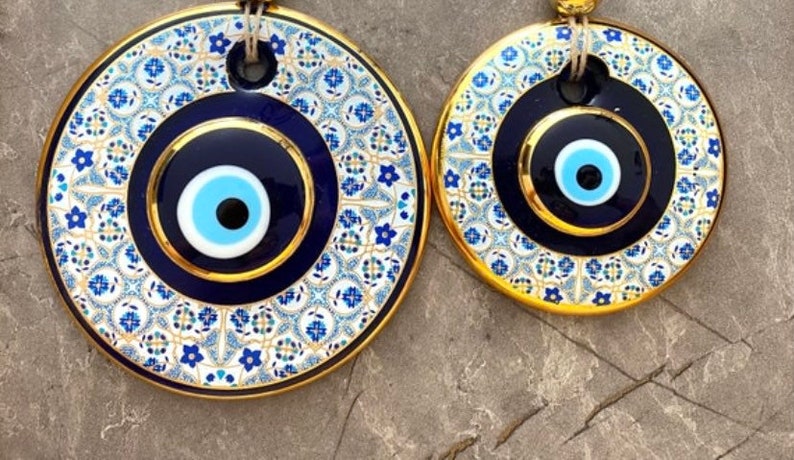 May include: Two round blue and white evil eye wall decor with gold trim. The larger evil eye is surrounded by a white and blue patterned design.