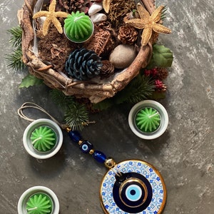 May include: A blue and white evil eye charm with gold accents hangs from a string of blue beads. The charm is surrounded by four small white bowls with green candles. The bowls are on a gray surface.