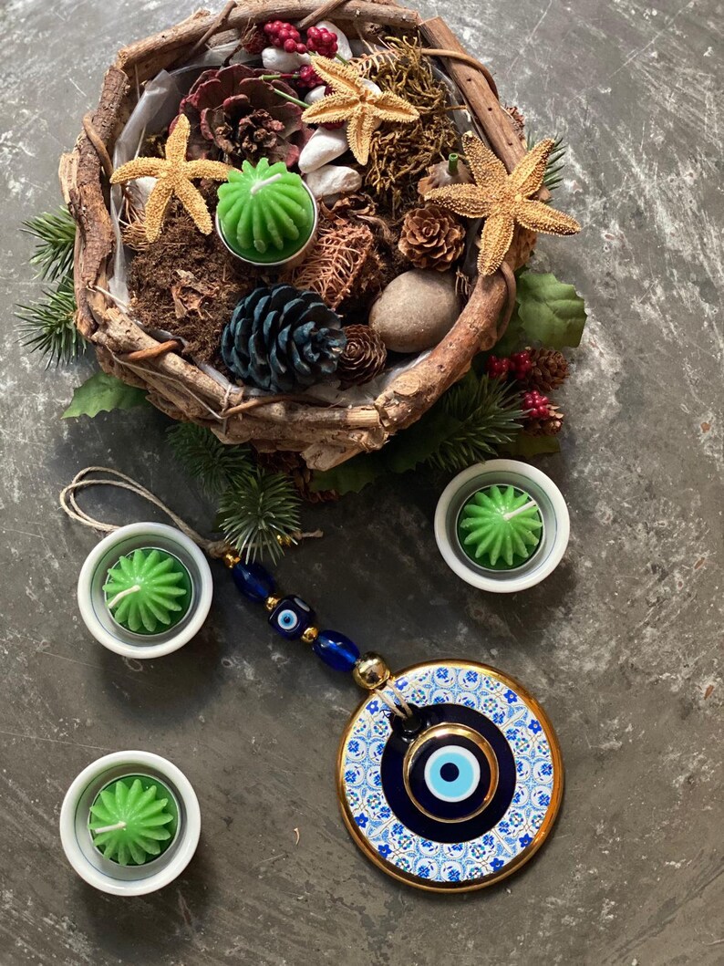 May include: A blue and white evil eye charm with gold accents hangs from a string of blue beads. The charm is surrounded by four small white bowls with green candles. The bowls are on a gray surface.