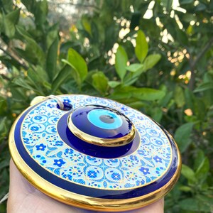 May include: A blue and gold evil eye wall hanging with a white and blue floral pattern. The evil eye is a symbol of protection against negative energy.