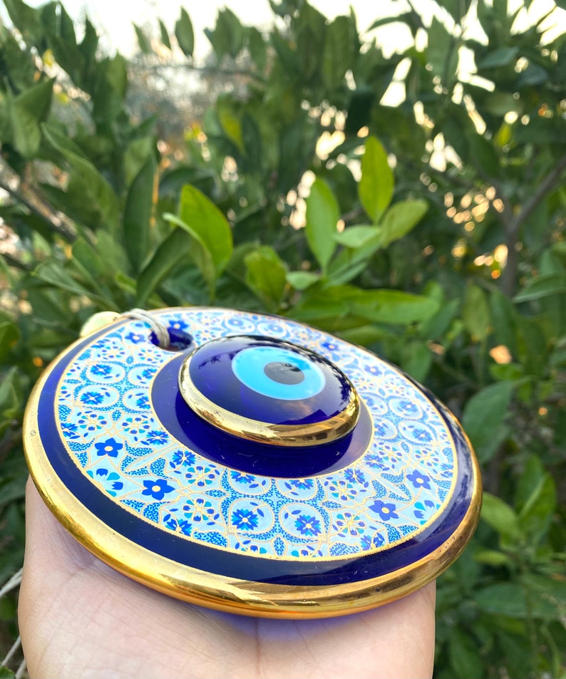May include: A blue and gold evil eye wall hanging with a white and blue floral pattern. The evil eye is a symbol of protection against negative energy.