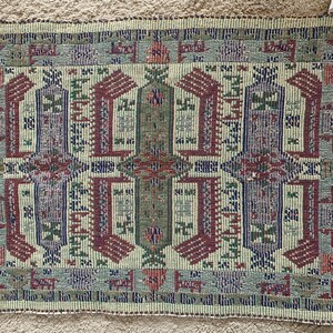 May include: A woven throw blanket with a geometric pattern in shades of green, brown, and cream. The blanket has a fringe border.