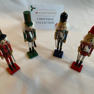 Op de afbeelding: Vier houten notenkraker-figuurtjes, elk met een ander ontwerp. De notenkrakers zijn geschilderd in rood, blauw, groen en goud. De notenkrakers staan op een wit oppervlak. De tekst "BOND STREET INTERIORS CHRISTMAS COLLECTION" is zichtbaar op de achtergrond.