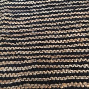 May include: A close-up of a woven rug with alternating black and beige stripes. The rug is made of natural fibers and has a textured surface.