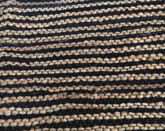 Puede incluir: Primer plano de una alfombra tejida con rayas negras y beige alternas. La alfombra está hecha de fibras naturales y tiene una superficie texturizada.