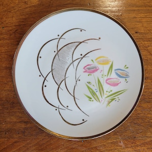 Op de afbeelding: Witte keramische plaat met een gouden rand en een bloemenontwerp. De plaat heeft roze, gele en blauwe bloemen met groene bladeren. Aan de linkerkant bevindt zich een abstract ontwerp in goud en grijs.