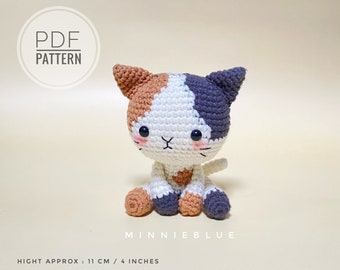 May include: A crocheted calico cat amigurumi toy with black eyes and a pink nose. The cat is sitting with its legs crossed and its tail curled around its body. The cat is approximately 11 cm / 4 inches tall. The image includes the text "PDF PATTERN" and "MINNIEBLUE".