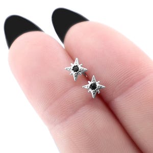 Può includere: Un paio di orecchini a lobo color argento, ciascuno caratterizzato da un design a stella a quattro punte con una piccola pietra nera rotonda incastonata al centro. Le montature in metallo a forma di stella hanno una finitura lucida e raggi.