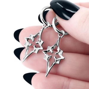 Può includere: Un paio di orecchini color argento. Ogni orecchino presenta un design traforato stilizzato a forma di stella con bordi appuntiti. Gli orecchini sono tenuti da una mano con smalto nero.