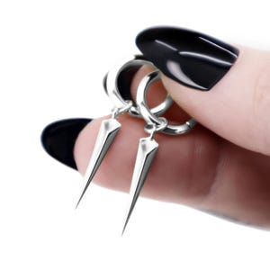 Op de afbeelding: Een paar zilveren oorringen met bungelende, puntige bedels. Elke oorbel bestaat uit een gepolijste ronde ring verbonden met een slanke, gefacetteerde metalen kegel die uitloopt in een scherpe punt, wat zorgt voor een strakke uitstraling.