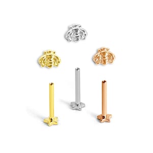 May include: A collection of three bee-shaped flat back studs in silver, gold, and rose gold tones. Each stud features a detailed bee top and a straight post with a star-shaped base, designed for body piercings in various metallic finishes.
