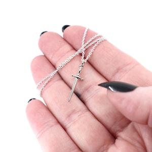 May include: A silver-toned necklace featuring a small, slender sword pendant suspended from a delicate, thin metal chain. The sword design includes a pointed blade, a straight crossguard, and a simple hilt, all crafted with a polished, reflective finish.